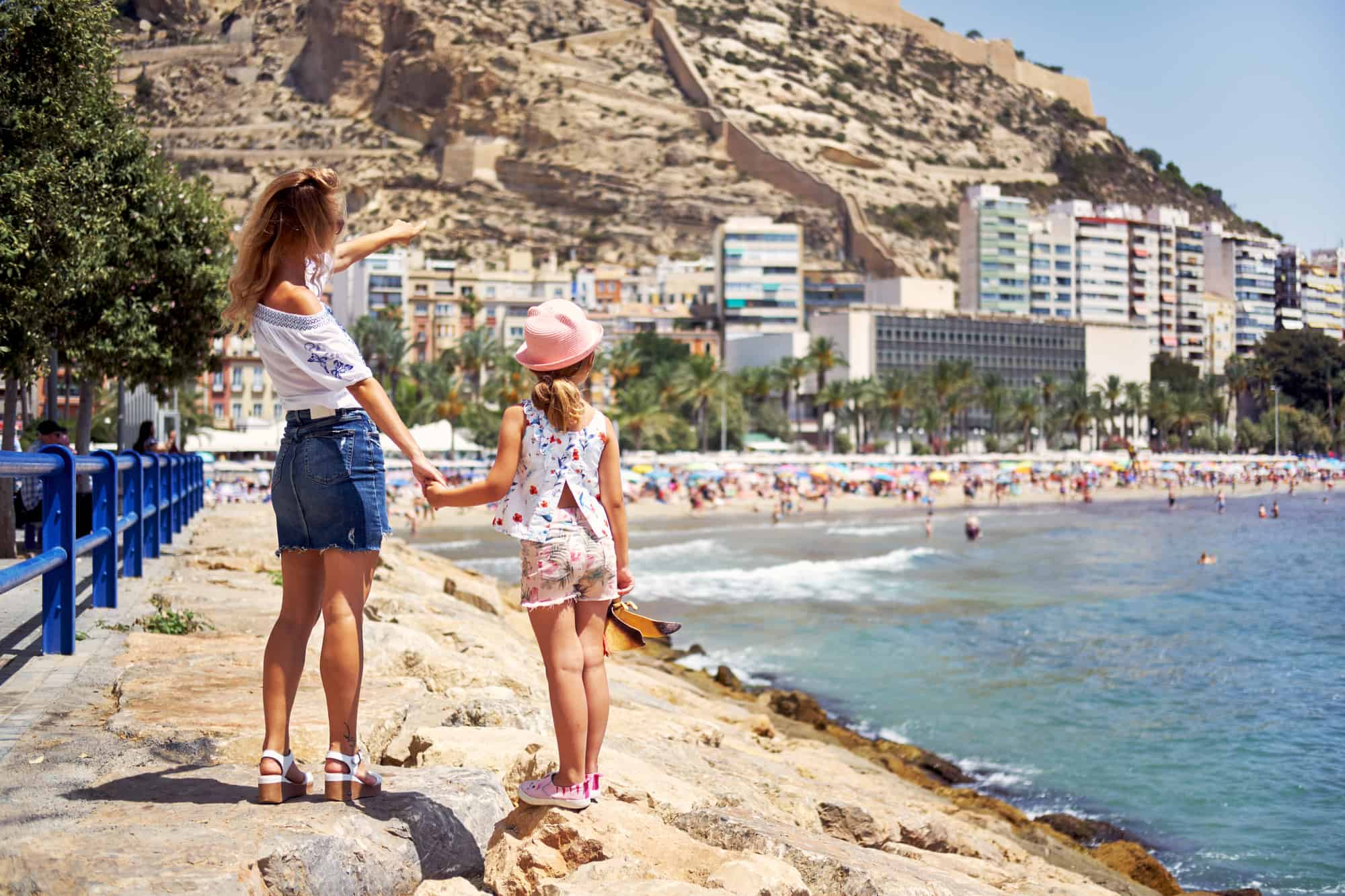 Mother and daughter on the Postiguet beach in summer, rear view. Mother showing to the Santa Barbara castle, fortification in the center, main landmark in Alicante city. Swimming and sunbathing tourist people in the background. Traveler, tourism, sightseeing and visiting new places concept. Costa Blanca. Spain