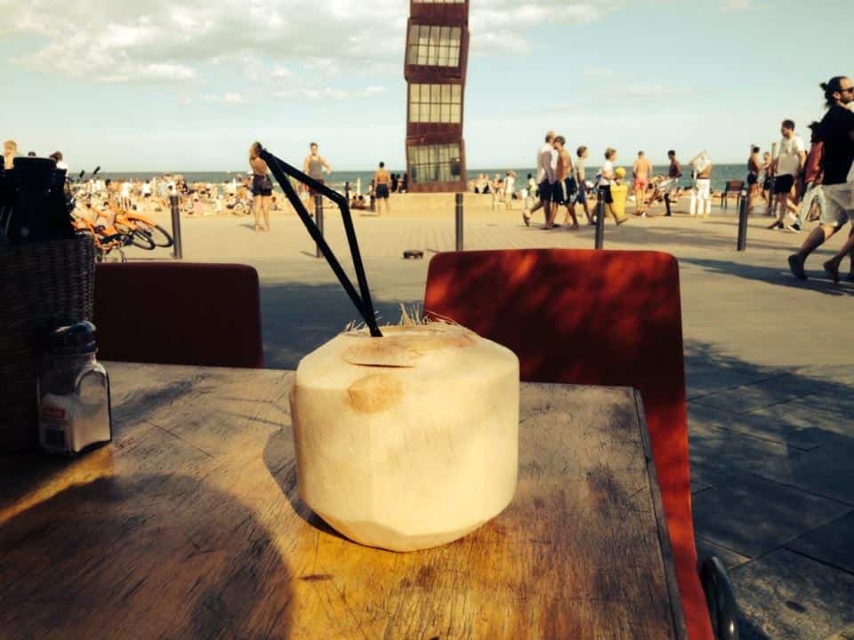 A refreshing coconut drink resting on the sandy beach of Barceloneta, Barcelona, evoking a tropical vibe and seaside relaxation.