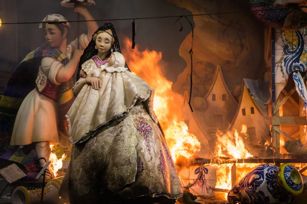 The image shows a "fallera" and a "falla" in fire during "fallas" in Valencia, Spain. This monuments are world heritage.