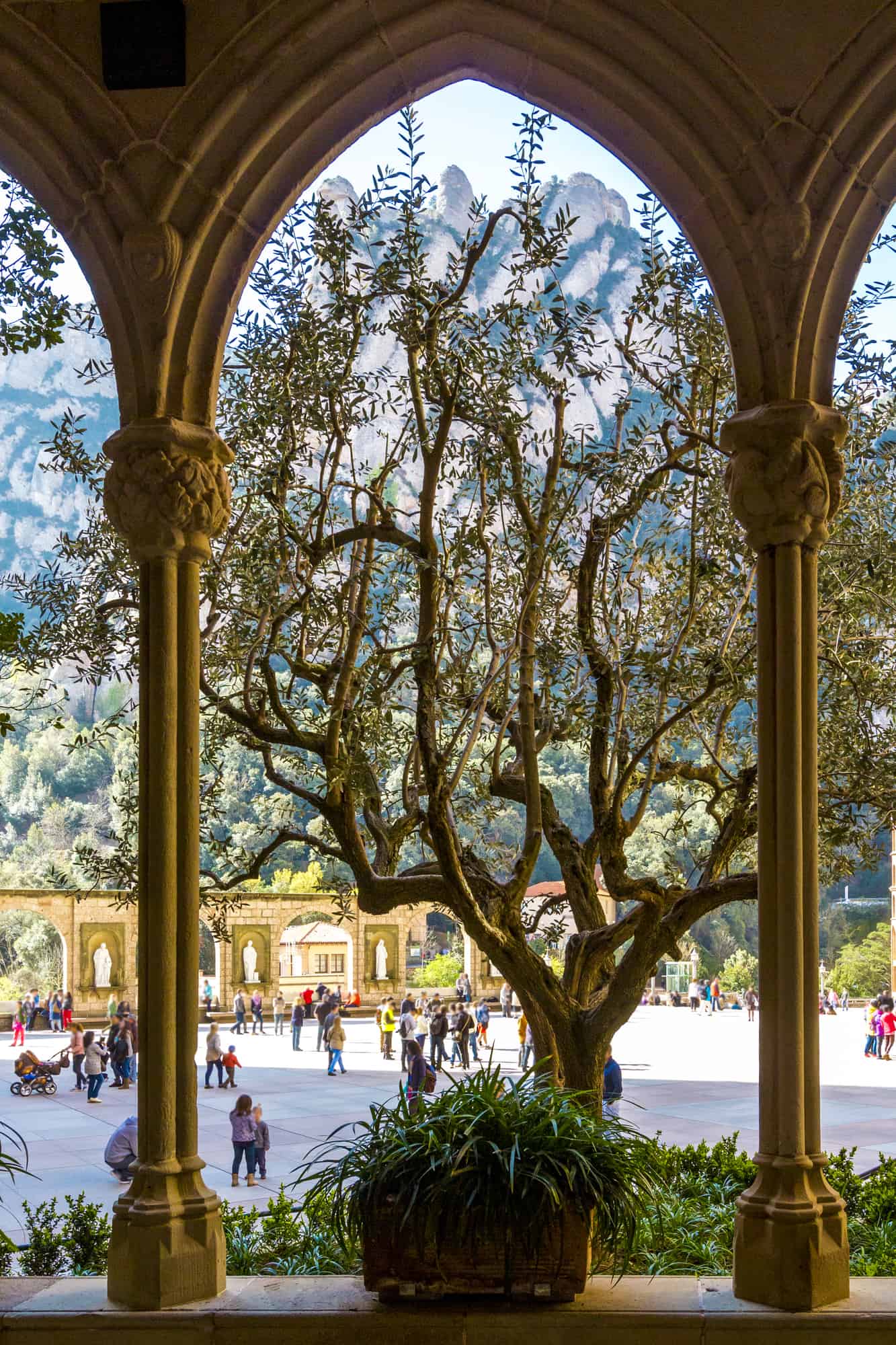 View of square, mountains and silhouette of tree through arch of Basilica Santa Maria de Montserrat Abbey, Catalonia, Spain