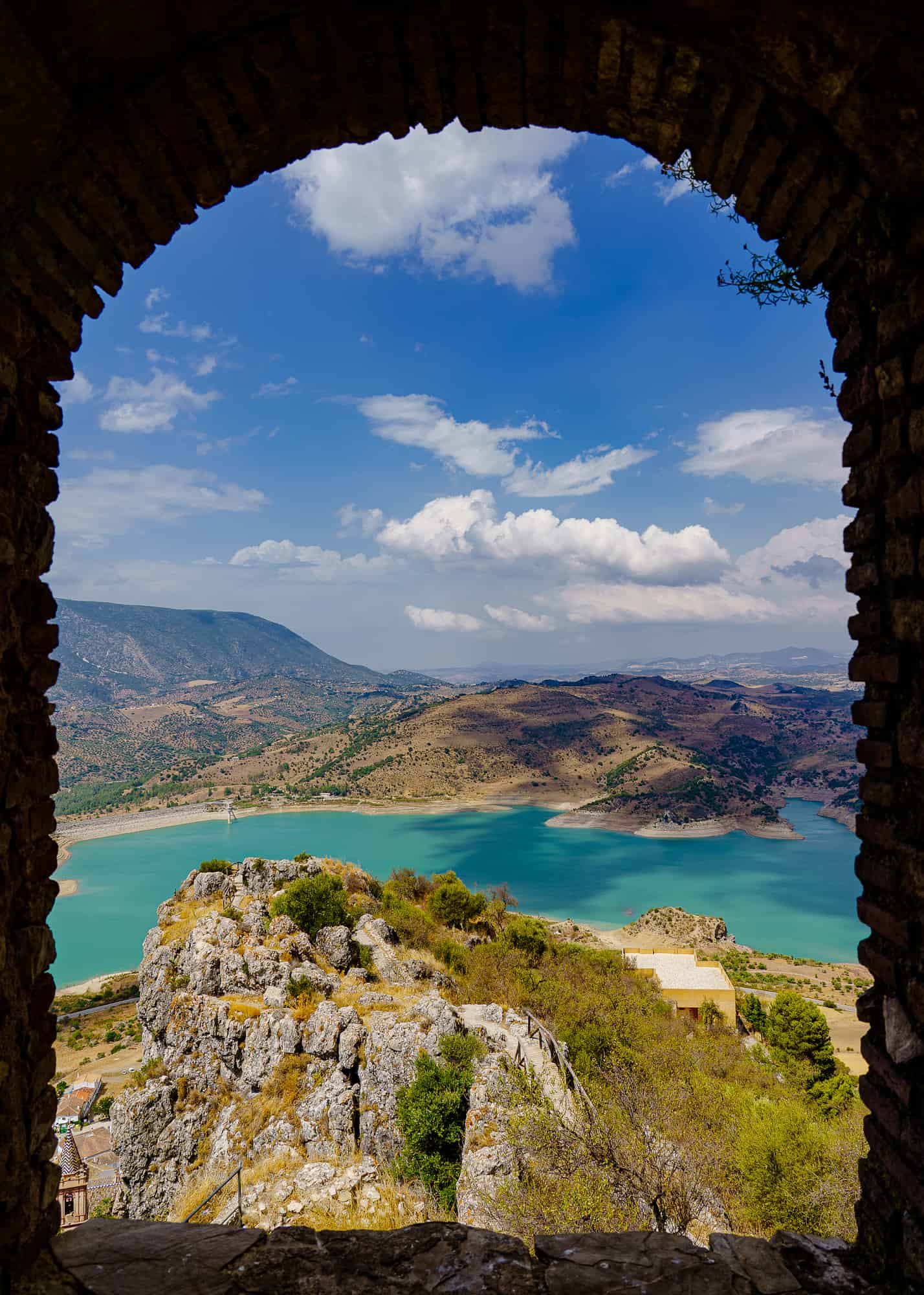 window framed view of turquoise lake, fields, road and mountains in the background