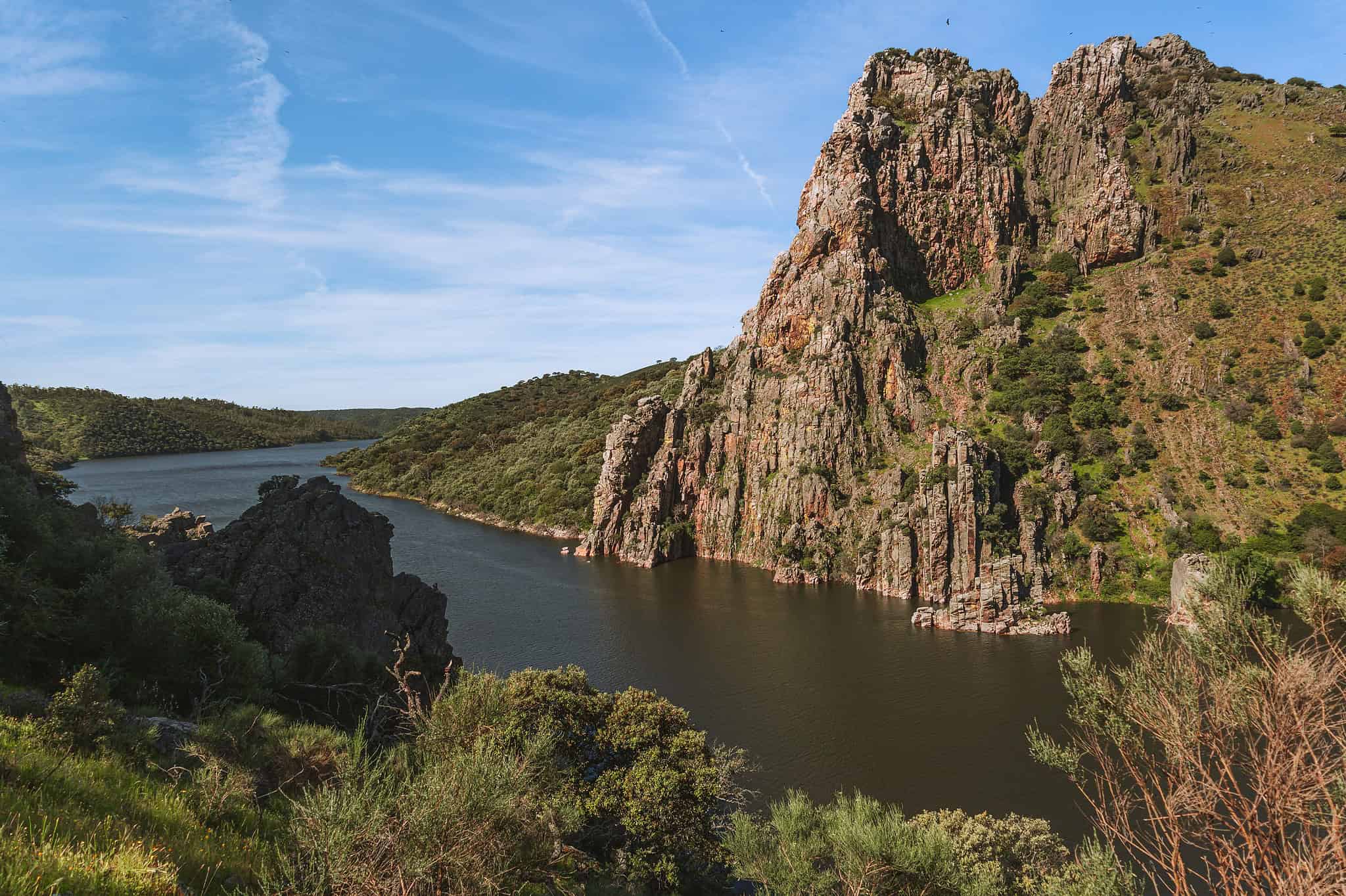 View of Peña Falcón rock formation from Salto del Gitano viewpoint in Monfragüe National Park on a sunny day, with the river visible in the background.