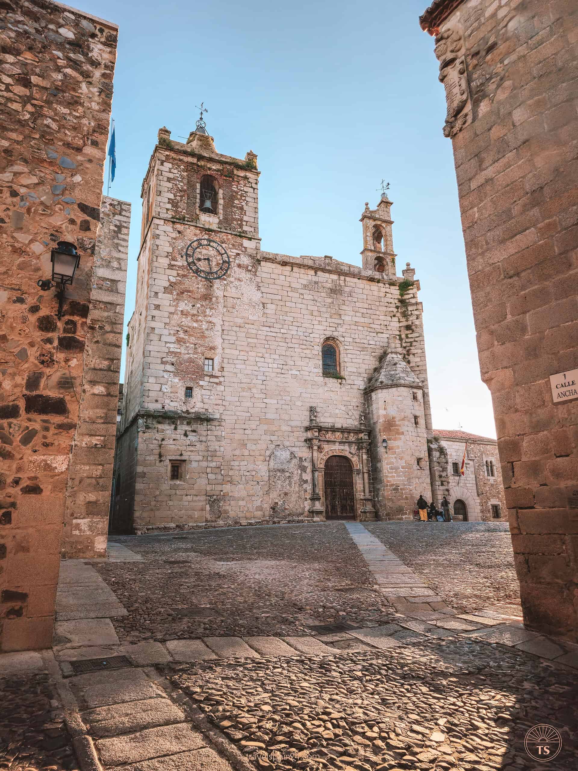Front view of Iglesia de San Mateo at Plaza de San Mateo in Cáceres, under a bright morning sky with the sun casting shadows along the historic walls.