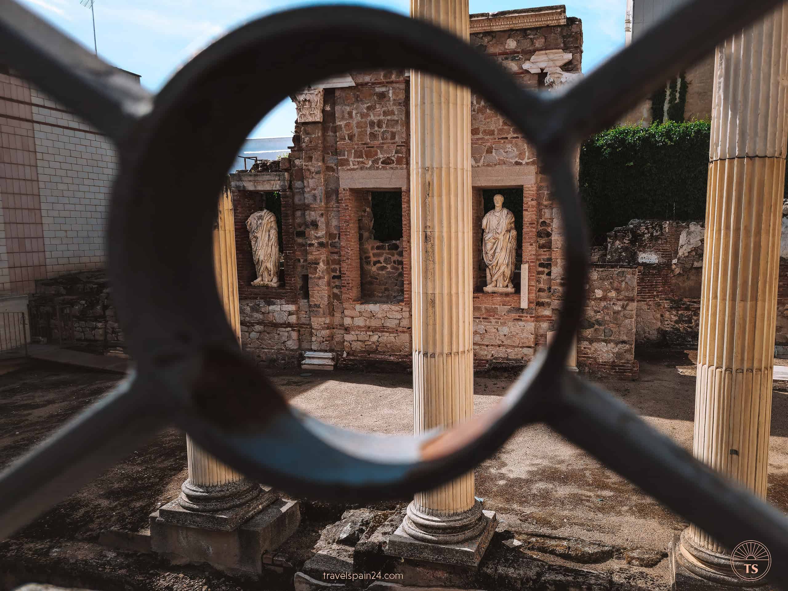Artistic view through a fence ring of the Portico del Foro Municipal de Augusta Emerita in Mérida, highlighting ancient pillars and statues from the Roman era