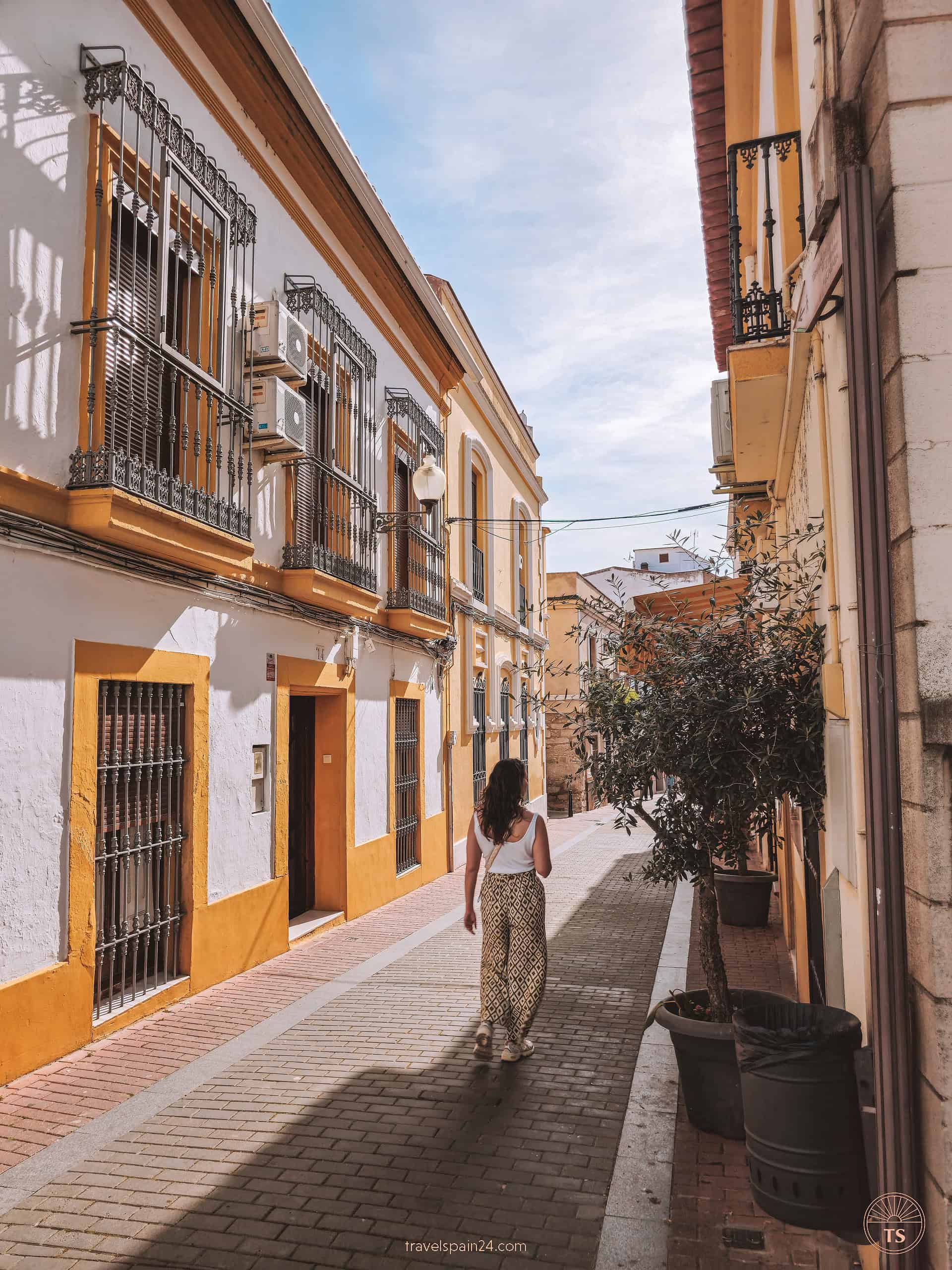 Filipa Ferreira strolling through the vibrant streets of Mérida, with yellow doors and balconies and olive trees lining the way, under a light blue sky, capturing the quaint charm of the city
