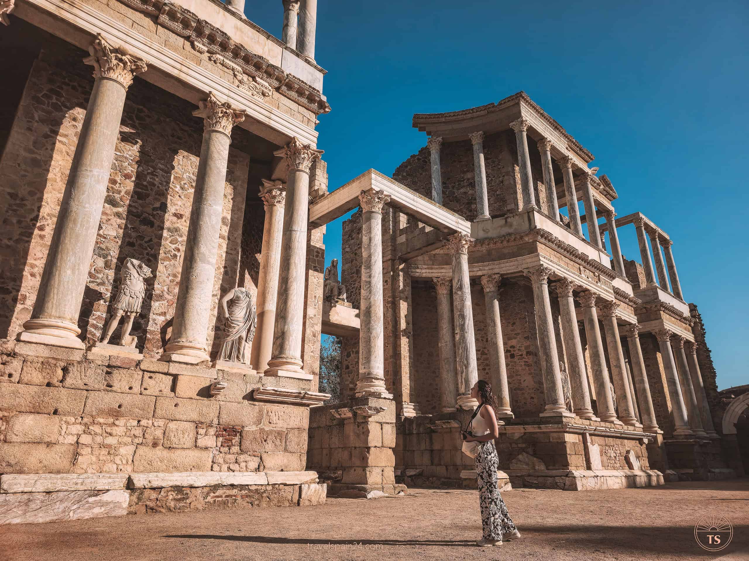 Filipa Ferreira in front of the Roman Theatre in Mérida, looking up at the impressive columns and ancient statues, enjoying the historical ambiance on a sunny day.