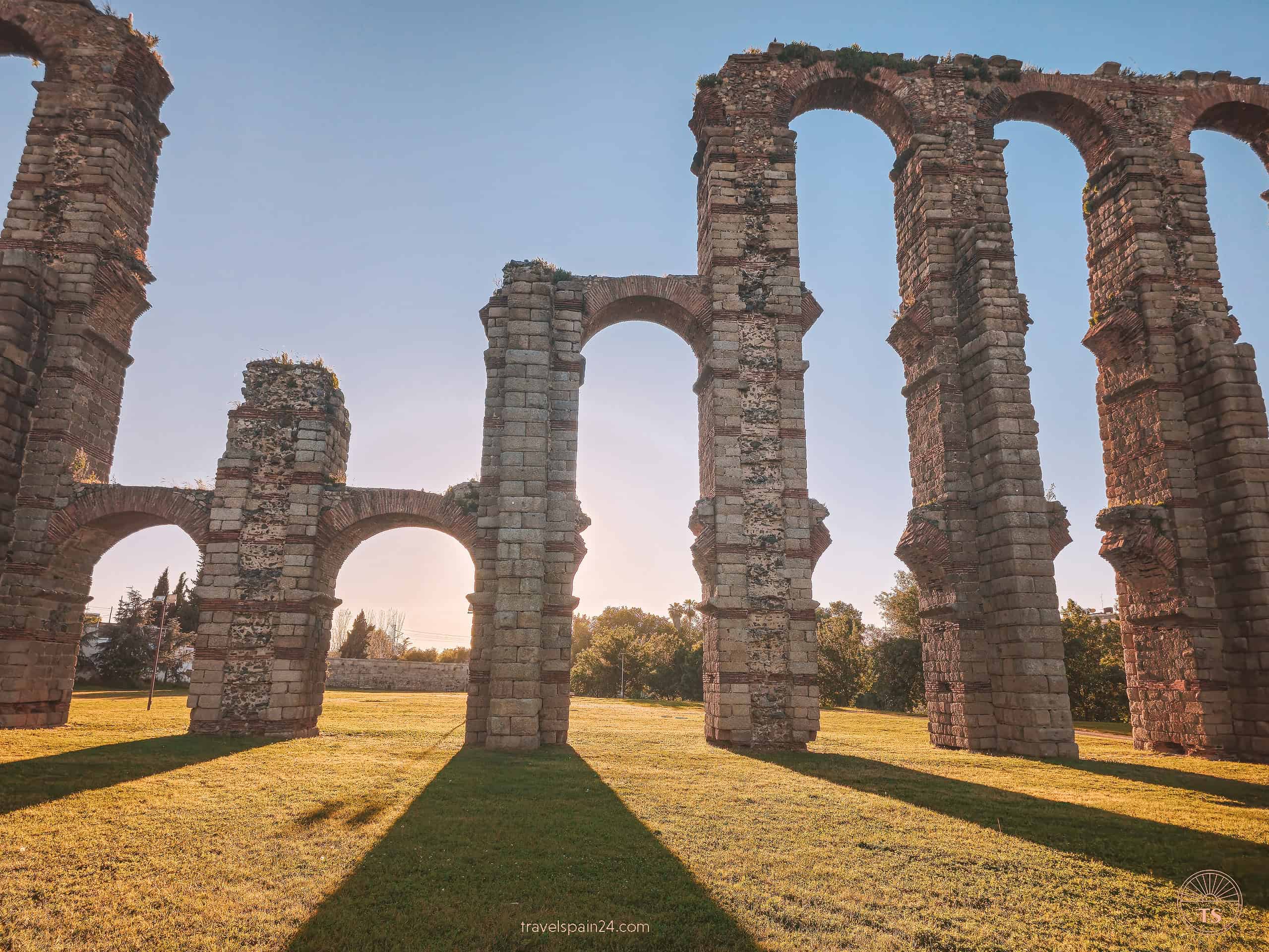 Sunset at Acueducto de los Milagros in Mérida, with the shadows of the aqueduct's pillars cast across the green grass and the sun setting behind, creating a stunning visual effect.