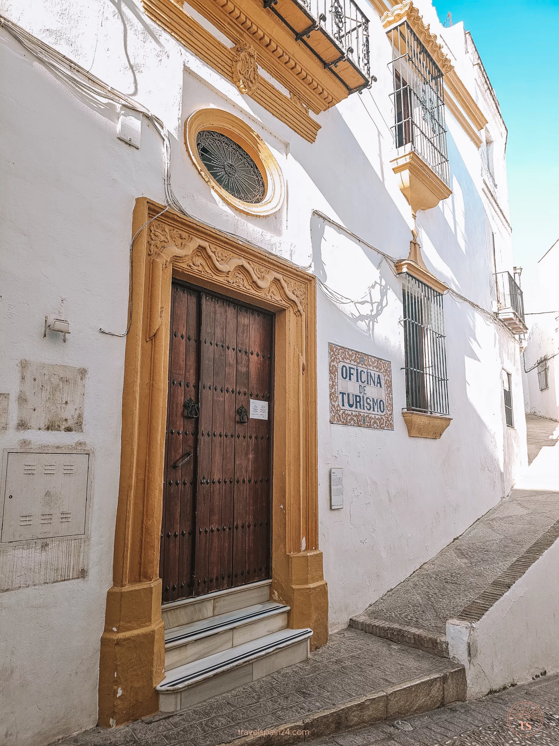 Tourism Office of Arcos de la Frontera. The entrance is a large wooden door surrounded by orange trim, set into white walls. 'Oficina de Turismo' is written on the wall.