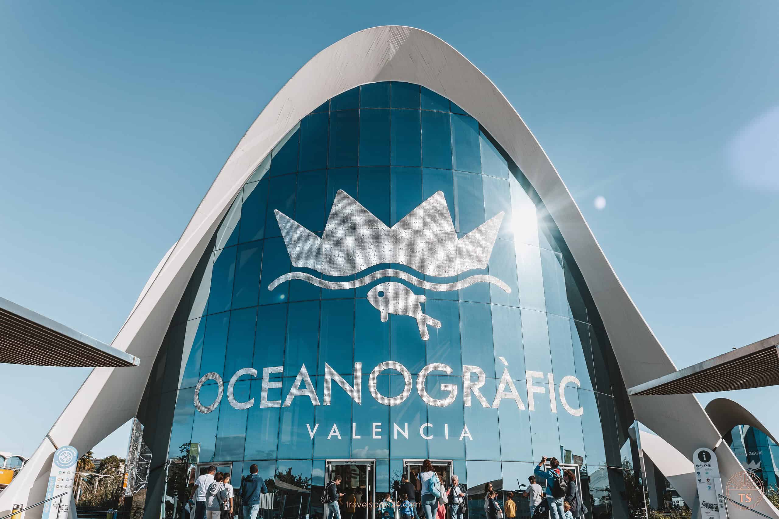 Busy entrance of Oceanografic Valencia, Europe's largest aquarium, featuring a striking architectural design with an arched entrance and large blue windows bearing the aquarium's logo.