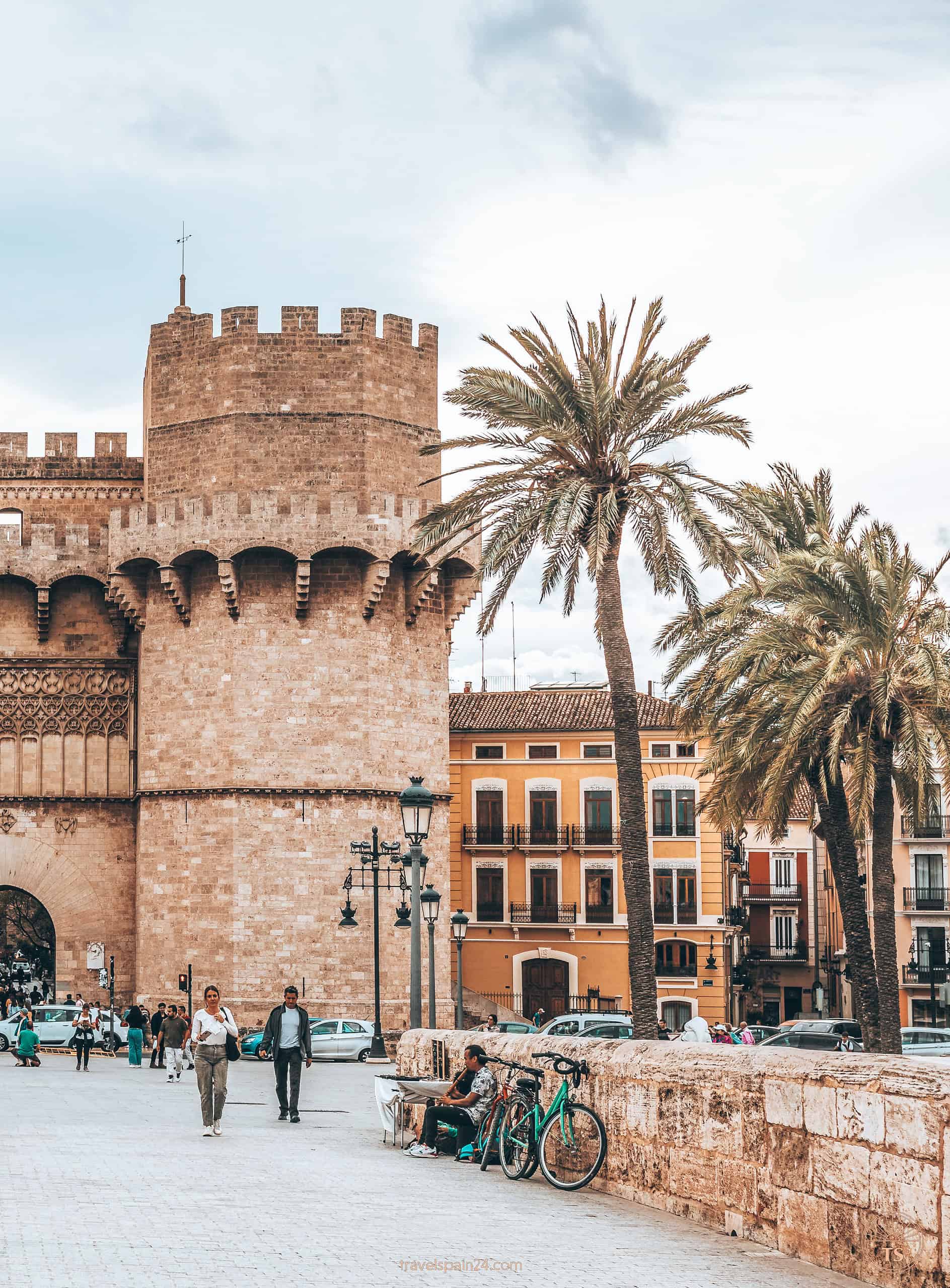 Close-up of the right tower of the Serranos Towers in Valencia, with people relaxing by the bridge wall, palm trees, passing cars, and light clouds setting a vibrant scene.