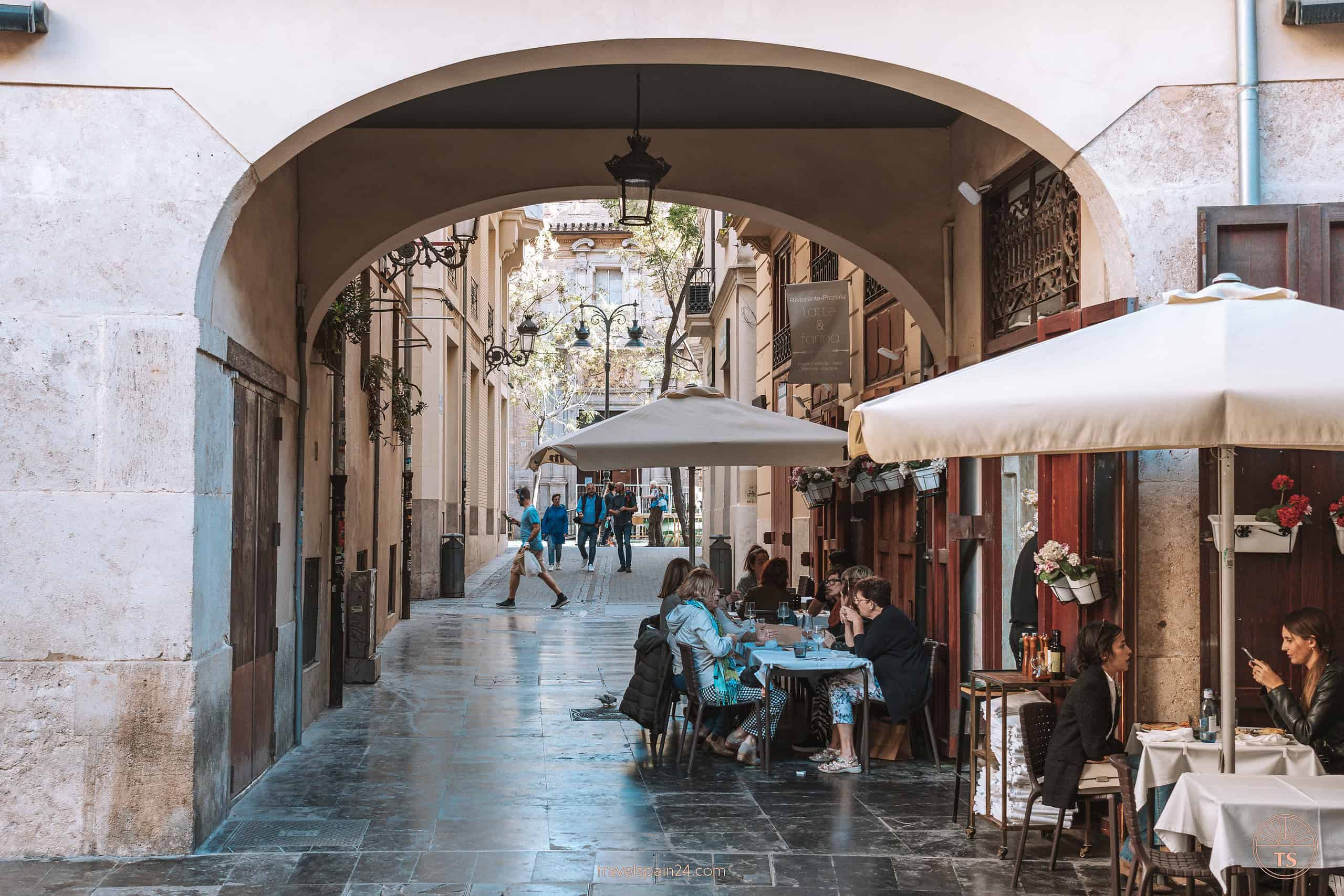 View of a bustling square in Valencia, with people enjoying time under an arched tunnel at café terraces, capturing the lively urban atmosphere.