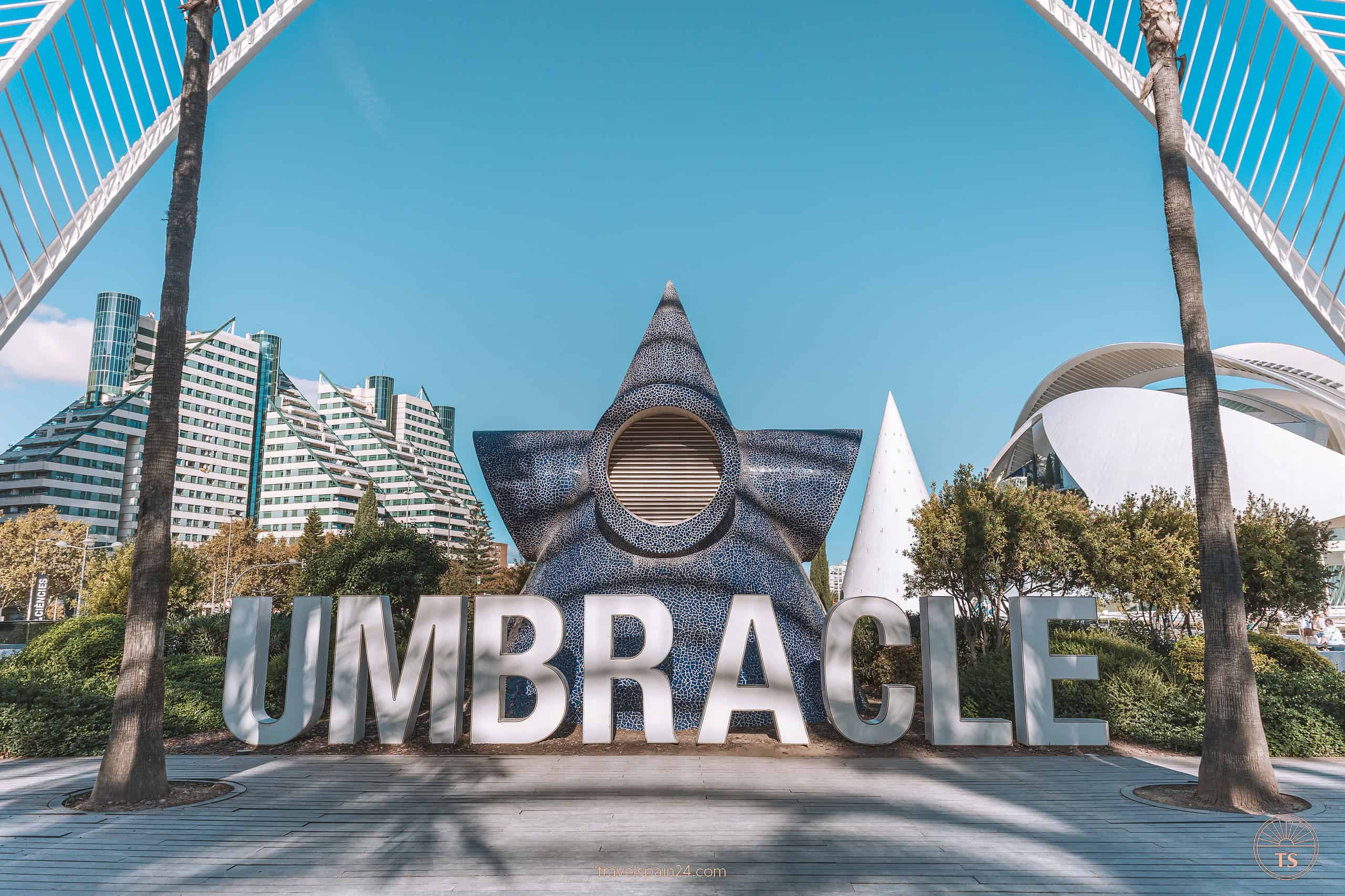 The large white UMBRACLE letters in L'umbracle Gardens, Valencia, with a backdrop featuring a large blue starfish sculpture and a glimpse of Palau de les Arts, highlighting the artistic flair of the area.