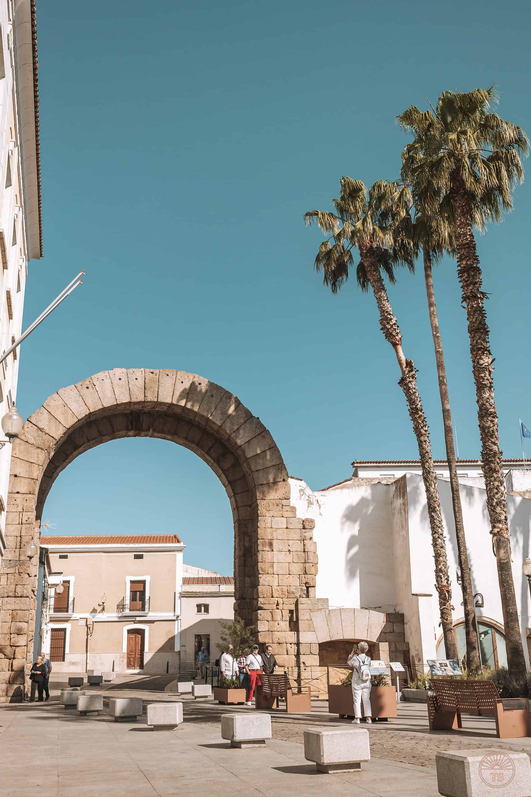 View of Trajan Arch in Mérida with people posing for photos, framed by palm trees, capturing a lively moment at this historic site.