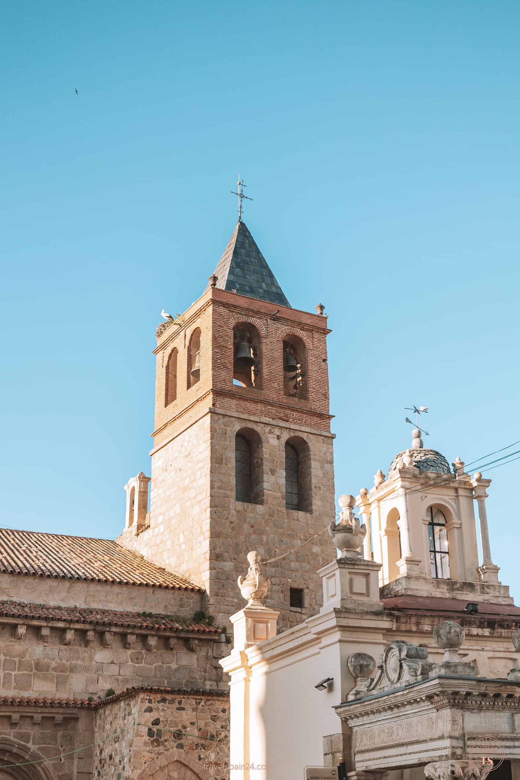 The tower of Basilica of Santa Eulalia in Mérida topped with a cross and a stork perched at the peak, with large church bells visible—a significant historical landmark in the city.