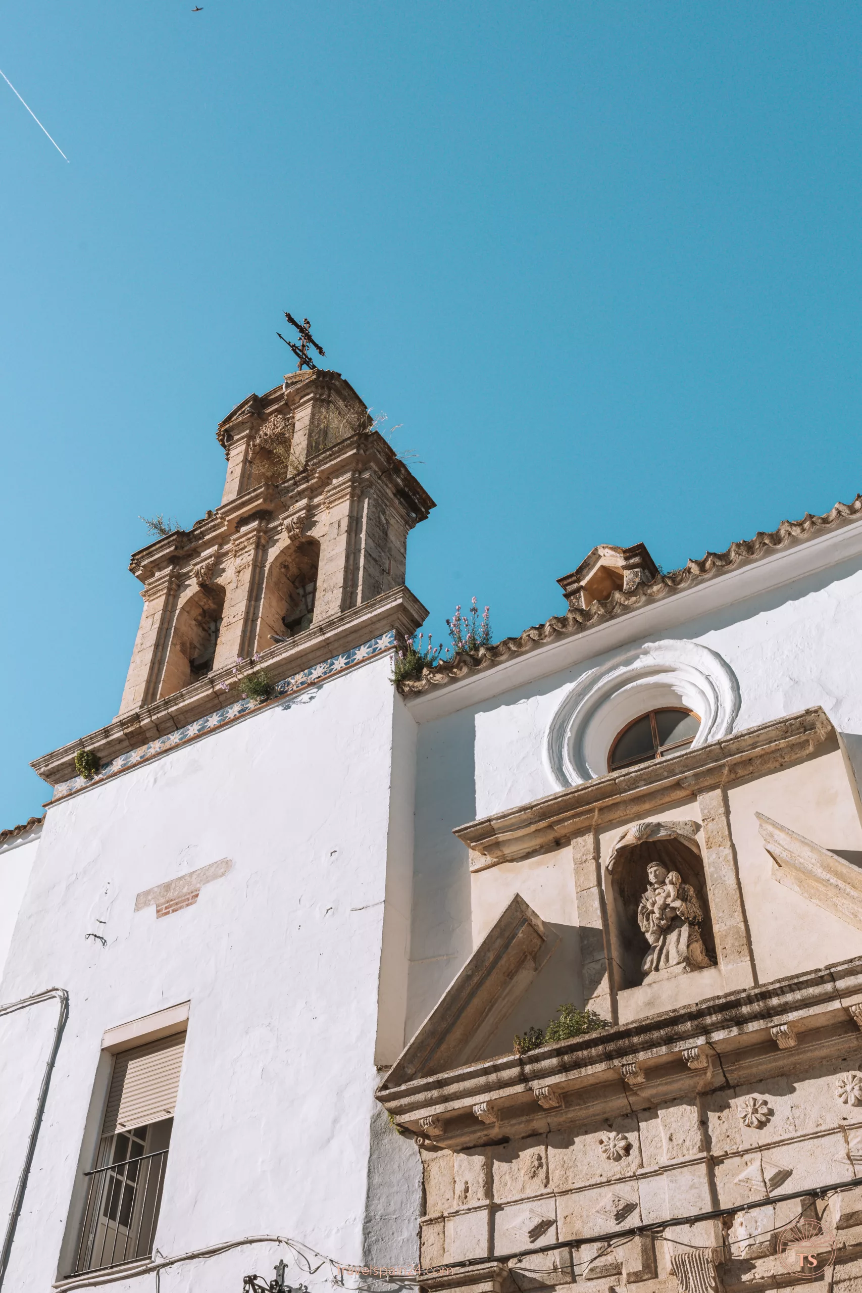 Iglesia de San Juan de Dios in Arcos de la Frontera features a small tower with a cross and tower clocks, and a sacred statue embedded in the wall.