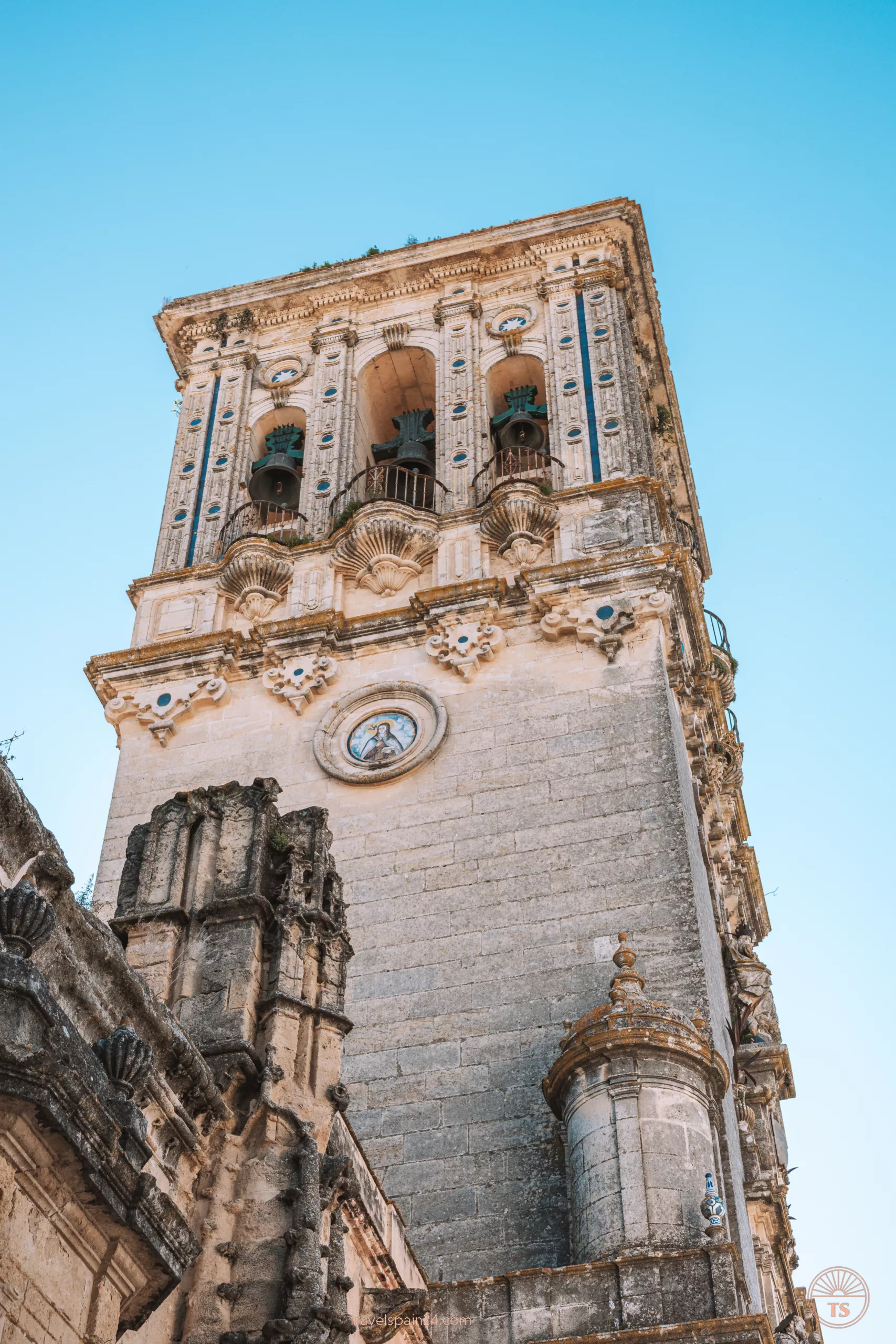 The tower with clocks of the Basilica Menor de Santa María de la Asunción in Arcos de la Frontera, an old Gothic church. The tower has a round tile depicting Mary and intricate architectural carvings.