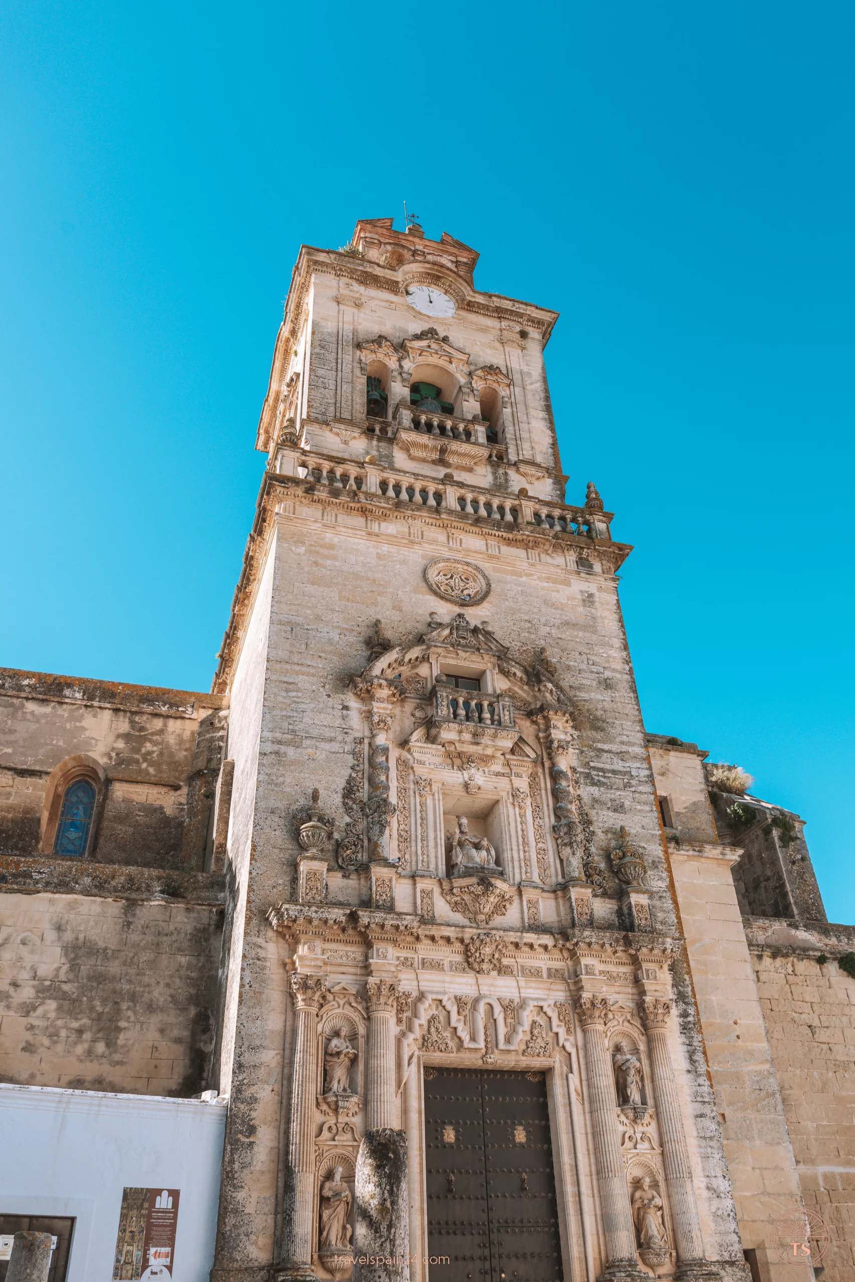Entrance and clock tower of Iglesia de San Pedro in Arcos de la Frontera. The old wooden doors are beneath a sacred carved image, and the tower features detailed architectural carvings.