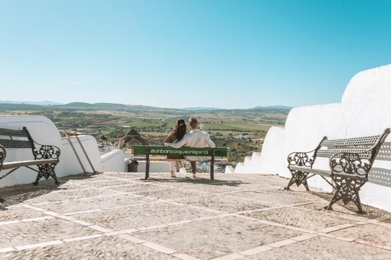 Timon van Basten and Filipa Ferreira sit on the famous bench with a panoramic view in Arcos de la Frontera, known as #unbancoqueinspira. It's a sunny day with a blue sky.
