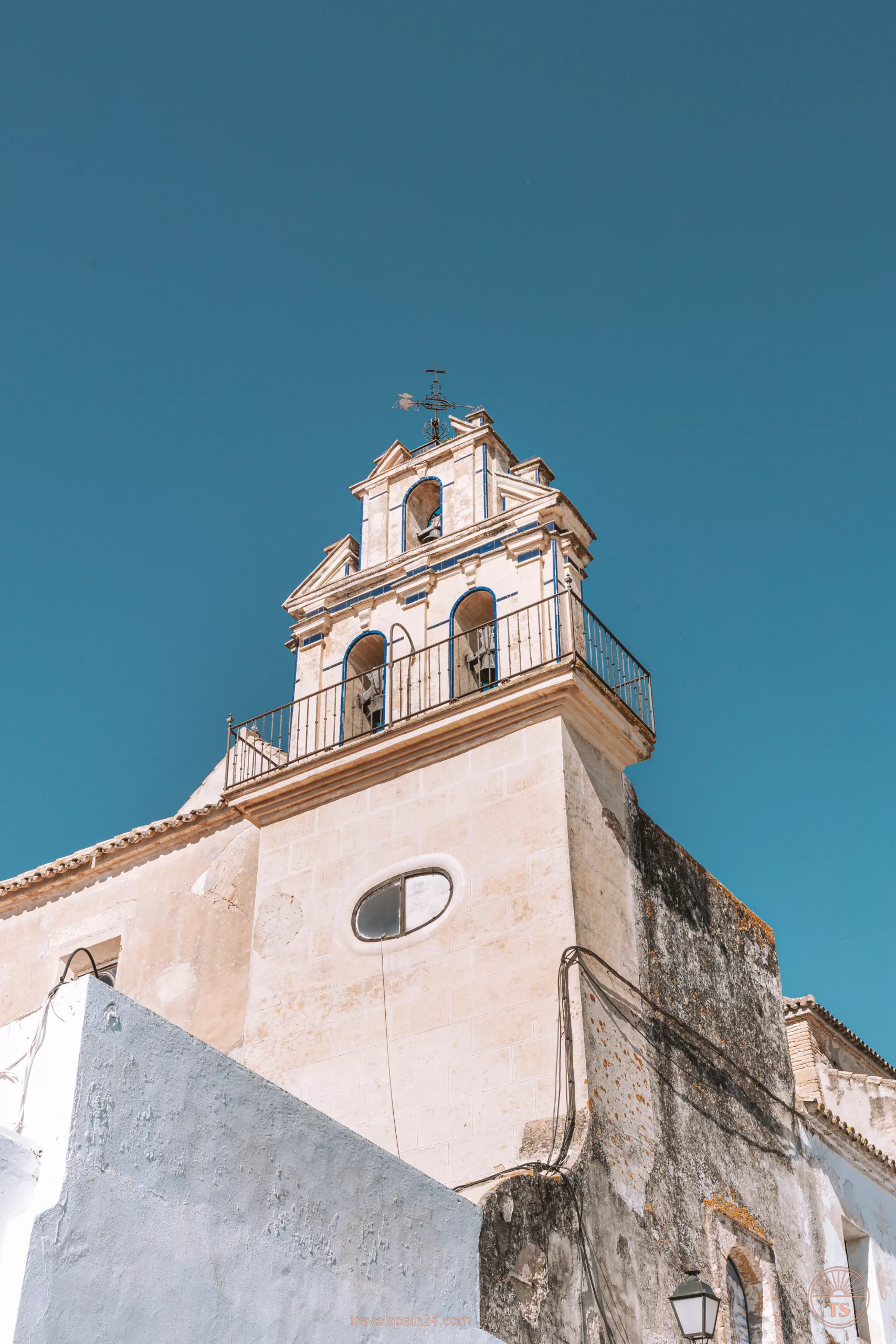 The tower of Iglesia de San Agustín in Arcos de la Frontera, set against a clear blue sky. The tower has three bells and a cross at the top.