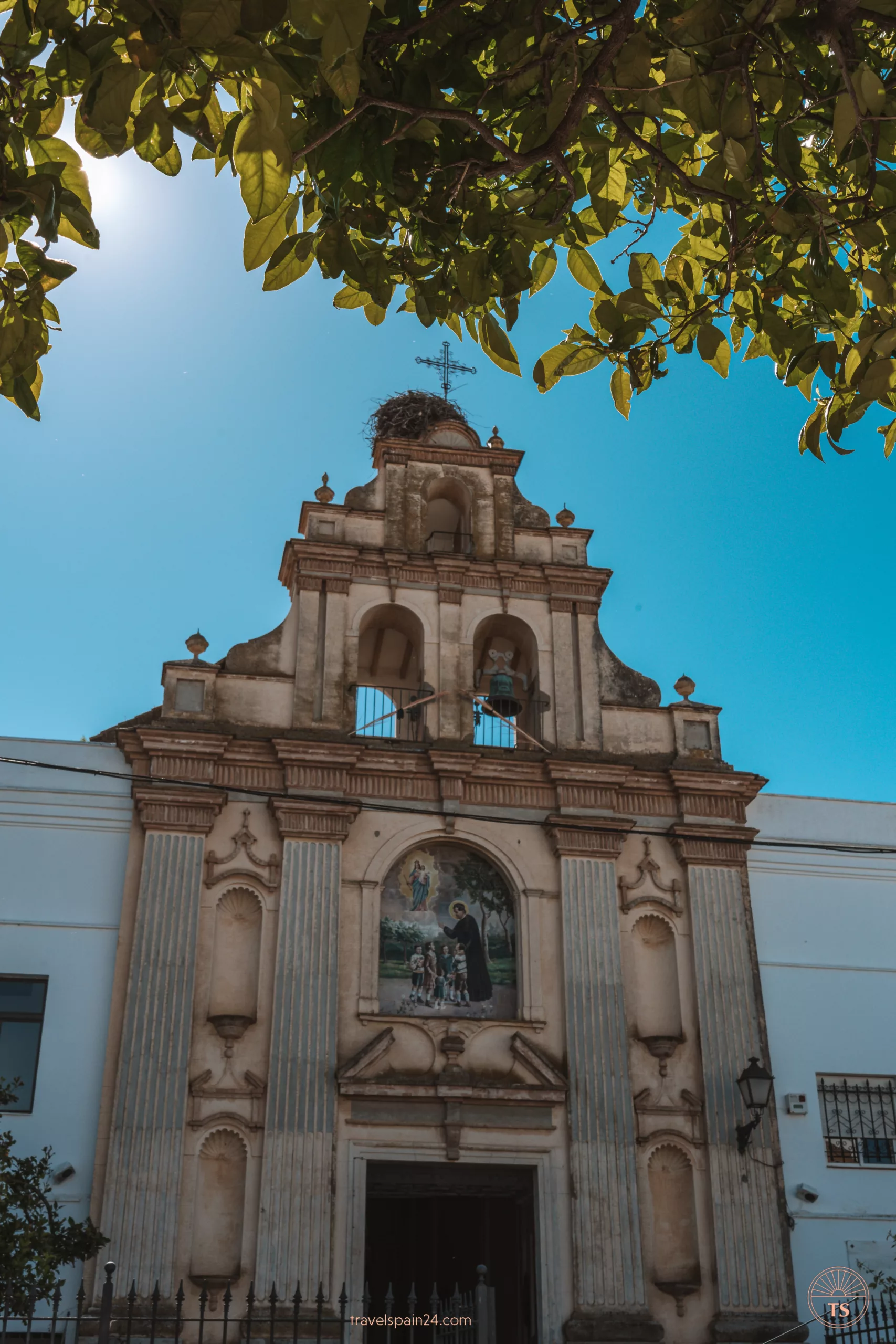 Iglesia de María Auxiliadora in Arcos de la Frontera, taken from under a tree. The tower has a cross and a stork's nest above three bells. An image of Jesus and a pastor watching over children is above the main entrance.