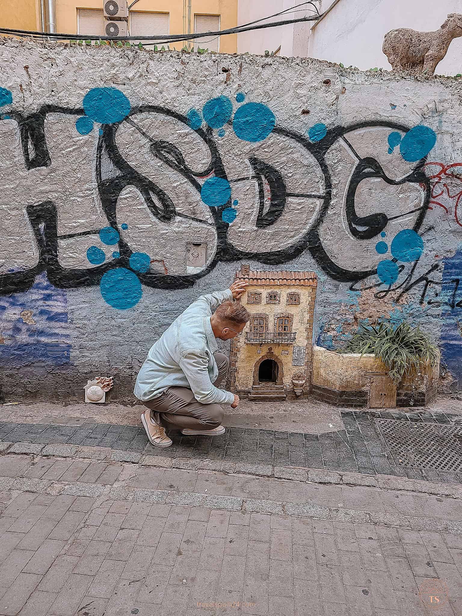 Timon van Basten crouching and peeking into The Cat House in Valencia, a charming little shelter built into the wall for street cats, illustrating the city's care for its feline residents.